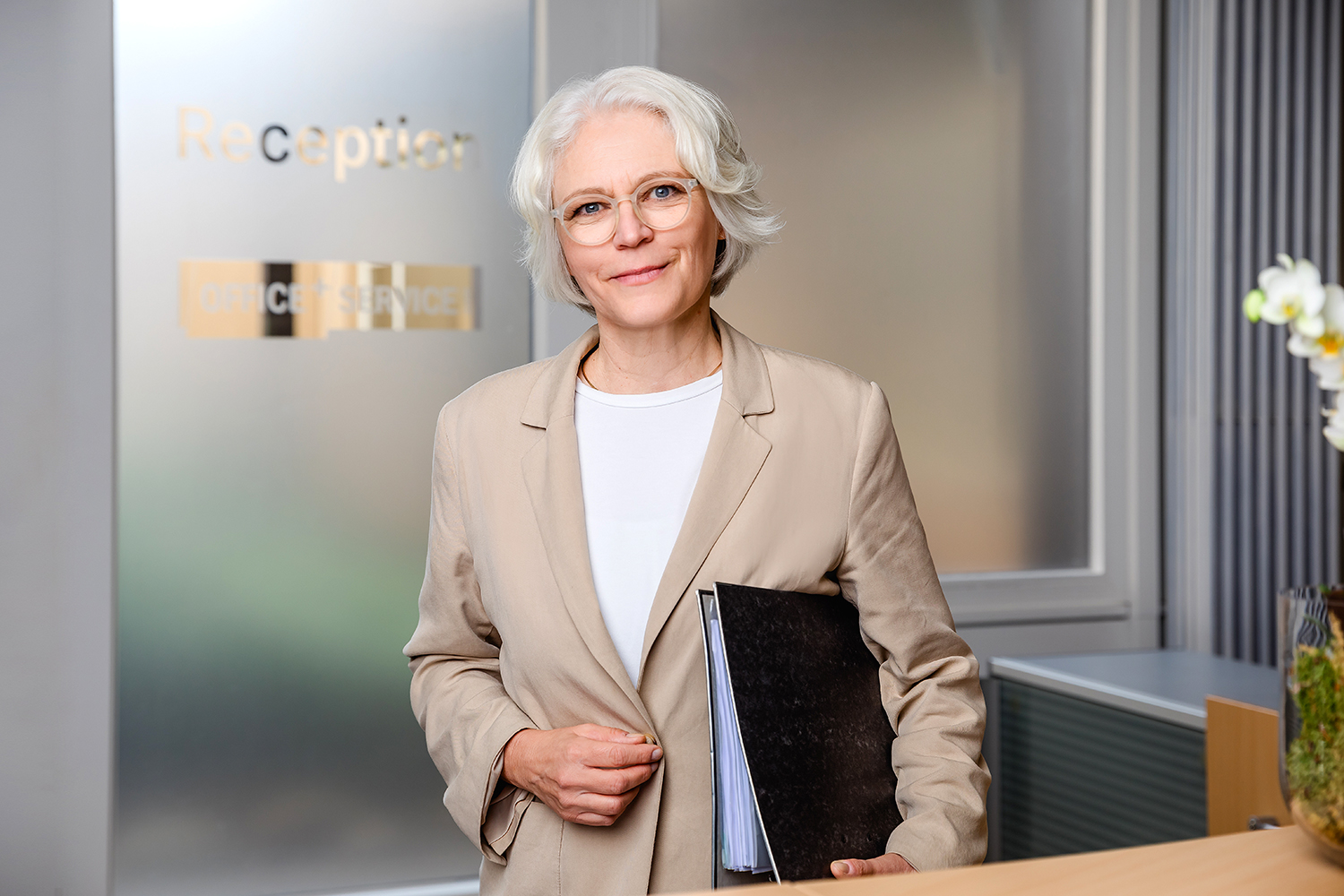 Portrait der Mitarbeiterin Barbara Derad, sie steht hinter der Rezeption mit einem Ordner unter dem Arm und schaut freundlich in die Kamera