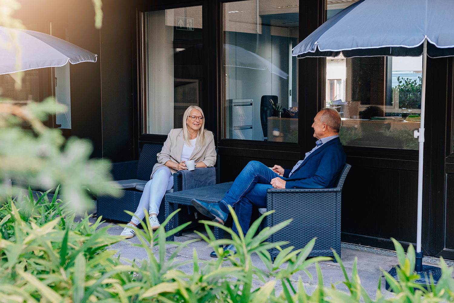 Office+Service-Außenbereich Zu sehen ist ein Außenbereich auf dem zwei Personen auf Gartenmöbel sitzen, mit Sonnenschirmen